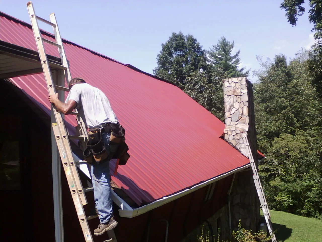 Roofer on a ladder beside a red metal roof and stone chimney — Soffit & Fascia Repair by Roofing Brothers in Sully